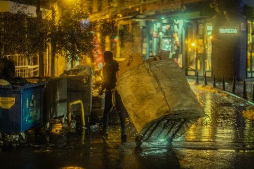 Worker moving a large bin of collected waste on a wet city street at night